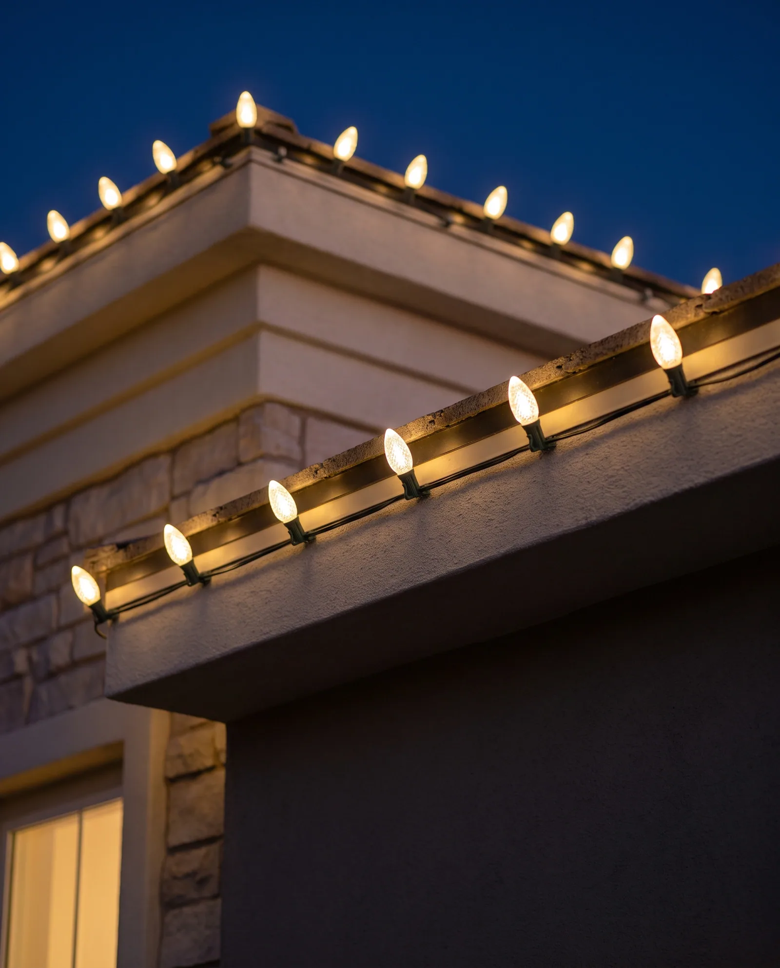 Close view of warm white C9 bulbs on socket wire clipped along a stucco and limestone eave at blue hour
