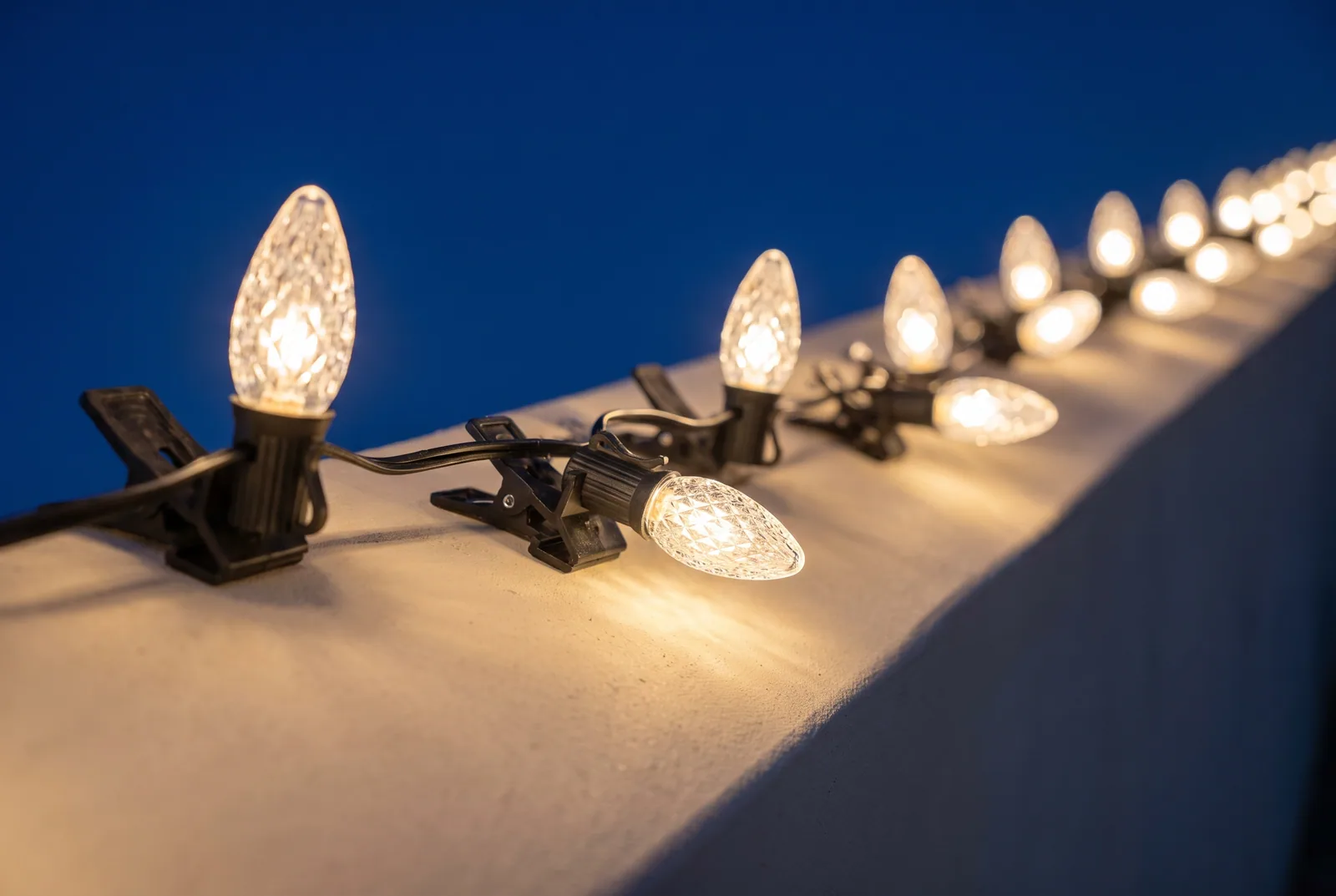 Warm white C9 bulbs on black socket wire with clips along a stucco parapet, the nearest bulbs sharp